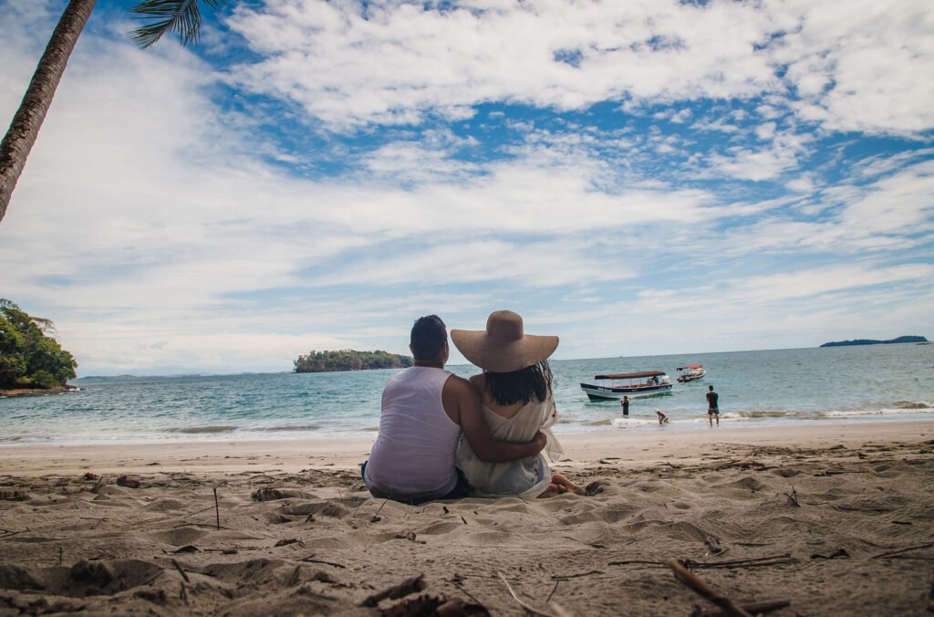 A horizontal shot of a couple sitting on a beach toward the calm blue water under the beautiful sky